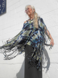 Person wearing a tie-dye poncho with fringe against a white wall