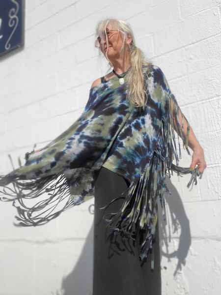Person wearing a tie-dye poncho with fringe against a white wall