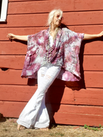 Person wearing a tie-dye shirt and white pants standing against a red wooden wall.