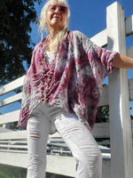 Person wearing a tie-dye shirt and white pants standing on a wooden deck with trees and blue sky in the background.