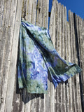A lightweight, semi-sheer scarf with a blue, purple, and green tie-dye pattern hanging from a rustic wooden fence against a clear blue sky.
