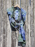 A blue and green tie-dye scarf draped over a woven hanger with a long beaded necklace, set against a weathered wooden plank background.