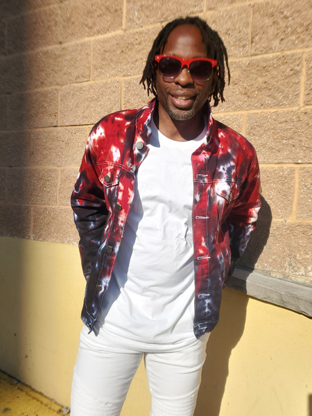 Person wearing a red and black tie-dye jacket, white shirt, and pants against a brick wall.