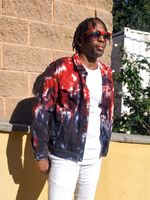 Person wearing a tie-dye jacket and sunglasses standing against a brick wall.