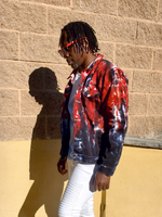 Person wearing a tie-dye jacket and sunglasses against a brick wall.