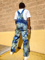 Person wearing blue tie-dye overalls standing against a beige wall. A back shot
