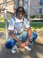Person wearing tie-dye overalls and sunglasses, sitting in front of a metal structure.