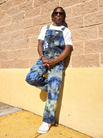 Person wearing tie-dye overalls standing against a beige wall.