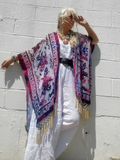 Woman wearing a kimono with floral patterns and fringe against a white wall.