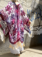 Person wearing a colorful patterned shawl with fringe against a textured wall.
