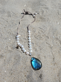 A beaded necklace with white freshwater pearls and a teardrop-shaped blue labradorite stone pendant, laid on a sandy beach.