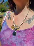 A close-up of a person wearing a necklace with white pearl-like beads, a dark metal chain, and a teardrop-shaped dark blue iridescent pendant, shown outdoors on a purple tie-dye top.