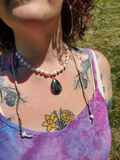 A close-up of a person outdoors wearing layered necklaces, including a pearl bead necklace with a dark teardrop pendant and a long silver-toned chain necklace with small beads.
