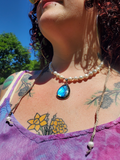 A close-up lifestyle shot of a woman outdoors wearing a beaded pearl necklace with a teardrop-shaped blue iridescent pendant and a long braided cord necklace.