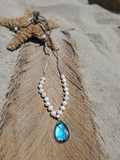 A beaded necklace with white pearl-like beads and a teardrop-shaped blue iridescent pendant, displayed on a large brown feather with a starfish prop on sand.