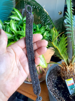 Hand holding a braided black leather cord bracelet with plants in the background. I'm showing the back side of the bracelet