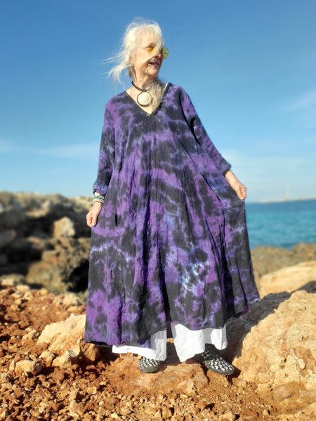 Woman in a purple tie-dye dress standing on rocks by the ocean.