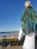A woman stands on a beach with her back to the camera, wearing a loose, oversized green and teal tie-dye top or poncho over a white dress. The background features a clear blue sky, ocean, and wooden grooved beams.