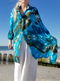 A woman with white hair stands on a beach wearing a flowing blue and teal tie-dye shirt jacket over a white top and white pants, with yellow-tinted sunglasses against a clear blue sky.