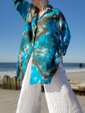A woman standing on a beach wearing a long-sleeve button-up shirt with a blue, teal, and olive tie-dye pattern, paired with white wide-leg pants.