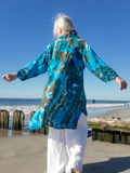 A woman standing on a beach with her back to the camera, wearing a long-sleeve tie-dye tunic top in vibrant blue, green, and brown, paired with white pants.