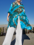 A woman wearing a vibrant turquoise and dark brown tie-dye button-up shirt with white wide-leg pants, standing outdoors on a sandy beach with a wooden fence and blue sky in the background.