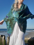 A woman on a beach wearing a flowing, semi-sheer poncho-style top in teal, green, and purple hues, paired with white wide-leg pants.
