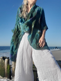 A woman standing on a beach wearing a loose, crinkled tie-dye shawl in teal, green, and blue over a white linen outfit, with the ocean and blue sky in the background.
