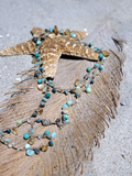 A handmade beaded necklace with turquoise, teal, and brown beads draped over a dried palm leaf on a sandy beach, styled with a starfish prop.
