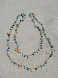 A double-strand beaded necklace made of turquoise, dark blue, and natural stone beads, laid out on a sandy beach.