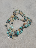 A multi-strand beaded necklace with turquoise, teal, and light brown beads on a gray sandy background.