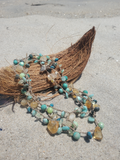 A bohemian-style beaded necklace with turquoise, green, and amber beads draped over a brown coconut shell on a sandy beach.