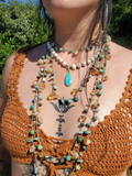 A close-up of a person outdoors wearing multiple layered bohemian-style beaded necklaces, including a turquoise pendant, over a rust-colored crochet top.