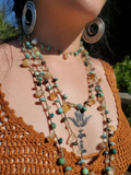 A close-up of a person wearing a bohemian multi-strand beaded necklace with turquoise, amber, and green beads, along with large silver spiral earrings and a brown crochet top.
