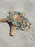A beaded necklace with turquoise, teal, and brown beads draped over a starfish on a sandy beach.