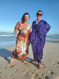 Two women in colorful dresses standing on a beach with ocean waves in the background.
