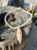 Necklace with an orange and white pendant on a piece of driftwood by the beach.