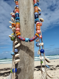 Colorful beaded necklace with natural elements hanging on a wooden post by the beach.