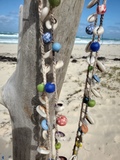 Decorative shell necklaces with colorful beads hanging on a wooden post by the beach.