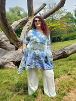 Woman wearing a tie-dye shirt and white pants standing in a natural setting with large logs and greenery.