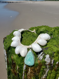 Necklace with white beads and a turquoise pendant on a mossy surface by a beach.