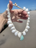 Hand holding a shell necklace with an aqua pendant on a beach.