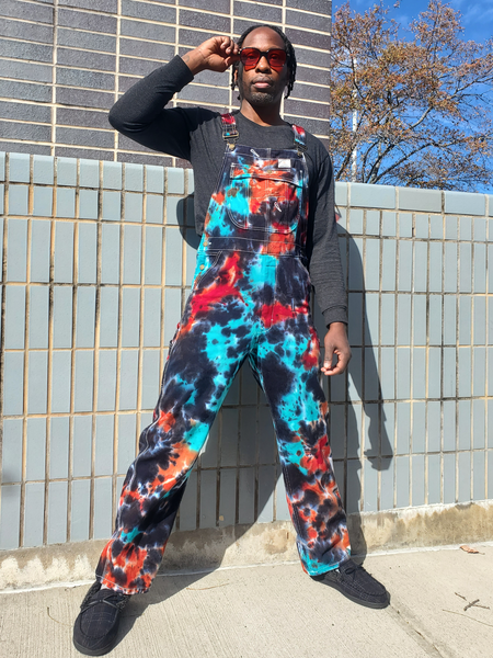 Person wearing colorful tie-dye overalls standing against a tiled wall.