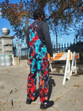 Person wearing a colorful tie-dye jumpsuit standing on a sidewalk with trees and a fence in the background.