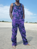 A man standing on a sandy beach wearing purple and blue tie-dye work overalls over a black tank top, with the sky in the background.