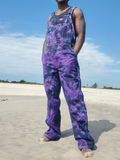 A person standing on a sandy beach wearing purple and black tie-dye work overalls over a black tank top, with a blue sky in the background.