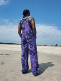 A man stands on a sandy beach from behind, wearing purple and black tie-dye overalls with a black tank top underneath, set against a blue sky.