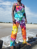 A person standing barefoot on a wooden pier at the beach wearing a sleeveless tie-dye jumpsuit with bright rainbow colors including blue, green, pink, purple, orange, yellow, and red. The background shows sand, ocean water, and a blue sky.