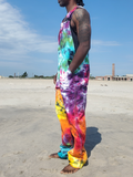 A man standing on a sandy beach wearing colorful tie-dye overalls with a rainbow pattern of pink, yellow, orange, green, blue, and purple. The background shows a clear blue sky and distant buildings.