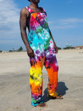 A person standing on a sandy beach wearing bright tie-dye overalls with a rainbow pattern of pink, blue, green, yellow, orange, and purple. The background shows a sandy beach, blue sky, and distant buildings.